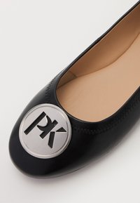 Ballerines noires avec un logo argenté rond arborant "PK" à l'avant. Texture en cuir lisse avec un détail de couture.