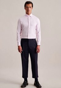 Jasnoróżowa koszula na guziki o dopasowanym kroju, w zestawieniu z ciemnoniebieskimi plisowanymi spodniami i czarnymi skórzanymi butami. Gładka tkanina, dopasowany fason.