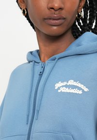 Sweatshirt azul com fecho éclair, feito de um tecido macio e texturizado. Apresenta um logótipo branco "New Balance Athletics" no peito e capuz ajustável com cordão.