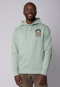 Lichtgroene hoodie met trekkoorden, voorzien van een rond grafisch ontwerp op de borst. Zacht materiaal met een voorzak en ribgebreide boorden.