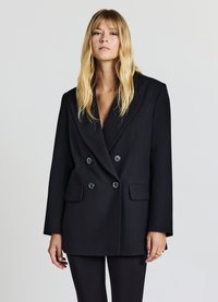 Blazer doppiopetto nero, vestibilità tailored, rever ampi, due tasche frontali, texture elegante e bottoni grandi che contrastano con uno sfondo chiaro.