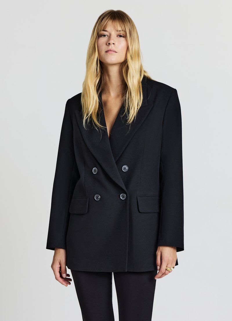 Blazer doppiopetto nero, vestibilità tailored, rever ampi, due tasche frontali, texture elegante e bottoni grandi che contrastano con uno sfondo chiaro.