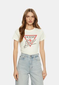 Weißes Baumwoll-T-Shirt mit einem roten dreieckigen Logo, dem Text "GUESS U.S.A." und silbernen Verzierungen, die "Love" buchstabieren. Kombiniert mit hellen Denim-Jeans.