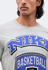 Νεαρό άτομο που φοράει ανοιχτό γκρι φούτερ Nike με μπλε και μαύρα γραφικά και κείμενο με θέμα το μπάσκετ.