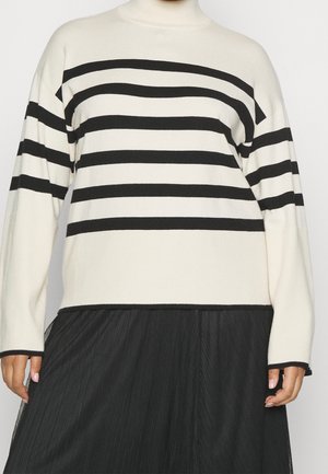 Pull col roulé blanc à rayures noires horizontales assorti d'une jupe noire plissée, montrant partiellement les mains et le torse.