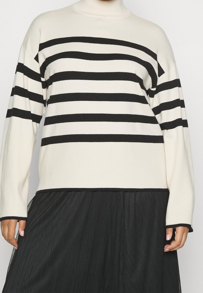 Pull col roulé blanc à rayures noires horizontales assorti d'une jupe noire plissée, montrant partiellement les mains et le torse.