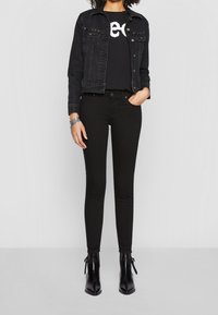 Chaqueta de mezclilla negra sobre una camiseta negra con un logo blanco, combinada con jeans skinny negros y botas negras hasta el tobillo con cordones.