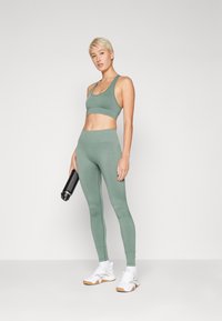 Soutien-gorge de sport vert menthe et leggings taille haute en tissu texturé, associés à des chaussures de sport à semelle blanche et gomme, tenant une bouteille d'eau noire.