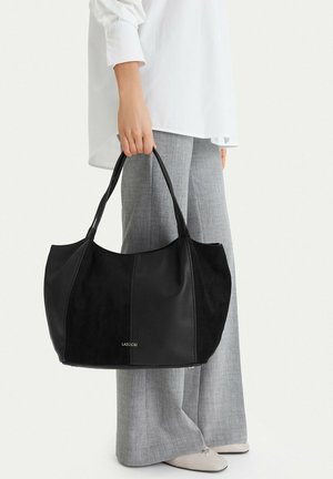 Persona con pantaloni grigi e camicia bianca che tiene una grande borsa nera con due manici e logo LASOCKI.