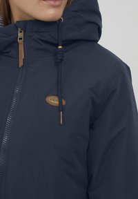 Giacca con cappuccio blu navy con zip frontale, dotata di una patch con logo, coulisse con dettagli dorati e un tessuto resistente e strutturato.