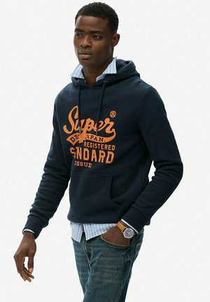 Marineblauwe hoodie met oranje tekst, kenguruzak en verstelbare capuchon. Draagt over een lichtblauw gestreept shirt en gecombineerd met jeans.