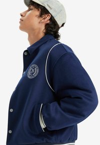 Navy blue wool jacket featuring a round logo patch, white piping accents, ribbed cuffs, and a button closure. Paired with a light denim cap.