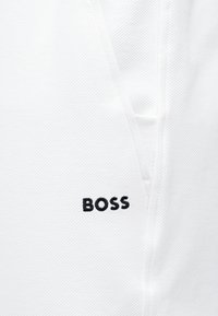 Κοντινή λήψη λευκού υφάσματος με κάθετη ραφή και τη λέξη "BOSS" κεντημένη με μαύρη κλωστή κοντά στη ραφή.