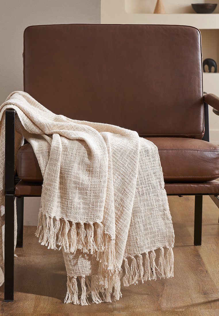 Coperta in cotone beige appoggiata su una sedia in pelle marrone, con bordi frangiati e una lavorazione testurizzata.