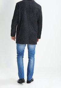 Abrigo texturizado gris oscuro con cuello, que llega a la mitad del muslo. Lleva puesto sobre jeans azul claro y zapatos negros. Vista trasera.