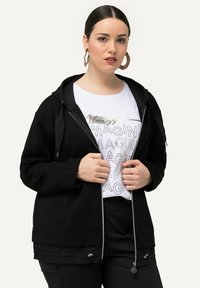 Haut de sweatshirt zippé noir avec une texture douce, doté d'une fermeture éclair à l'avant, de cordons assortis réglables et d'une coupe décontractée, associé à un t-shirt graphique blanc en dessous.