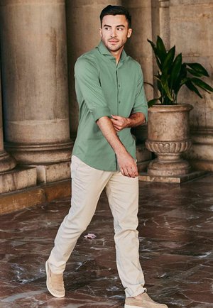 Man die de mouwen van een groen overhemd met knopen aanpast, lopend over een marmeren vloer in een gebouw met zuilen en een grote stenen plantenbak met een plant.