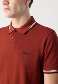 Červená polo tričko vyrobené z textúrovanej látky, s čierno-bielym prúžkovaným golierom a čiernym logom na ľavej strane. Trojgombíková léga.