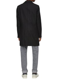 Langer schwarzer Wollmantel mit geradem Schnitt und Reverskragen. Kombiniert mit hellgrauen Hosen und weißen Sneakers.