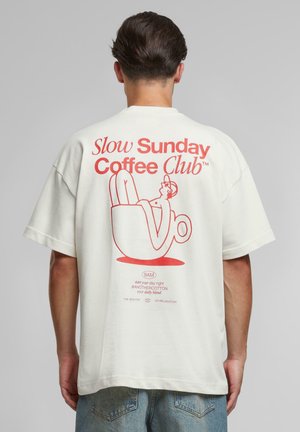 Bărbat purtând un tricou alb cu text roșu „Slow Sunday Coffee Club” și o ilustrație cu o persoană relaxându-se într-o ceașcă de cafea pe spate.