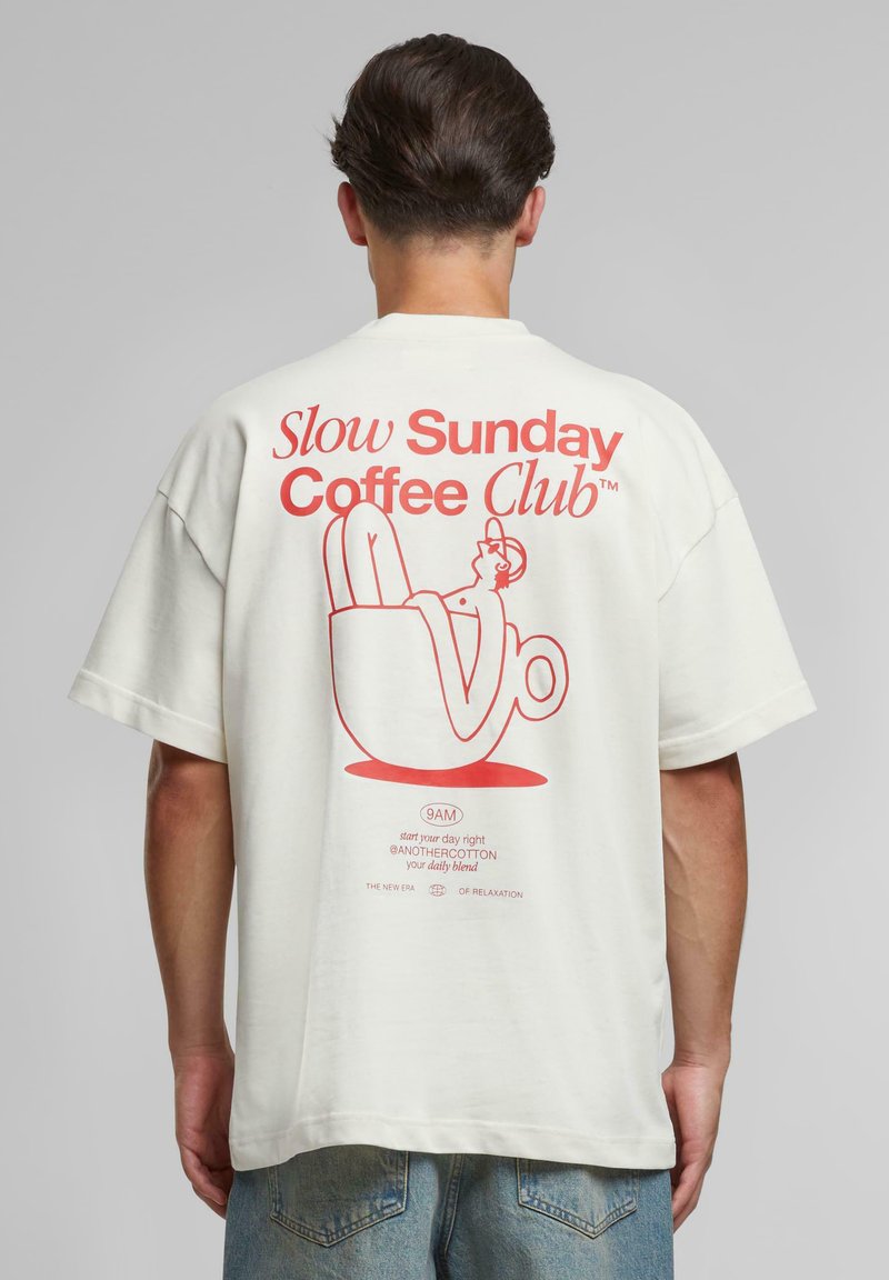 Moški nosi belo majico z rdečim napisom "Slow Sunday Coffee Club" in ilustracijo osebe, ki na hrbtu počiva v skodelici kave.