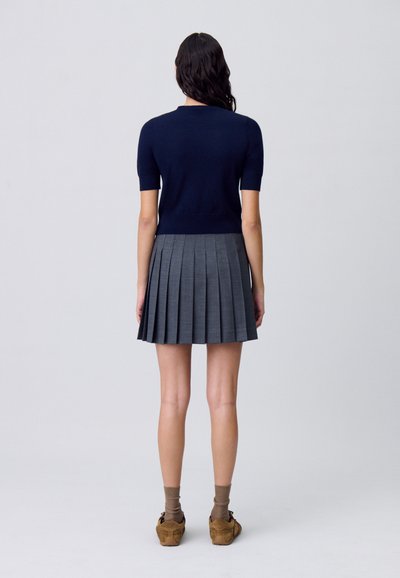 Top de punto de manga corta en color azul marino combinado con una falda plisada gris. Modelo con zapatos marrones y calcetines se encuentra de pie frente a un fondo liso.