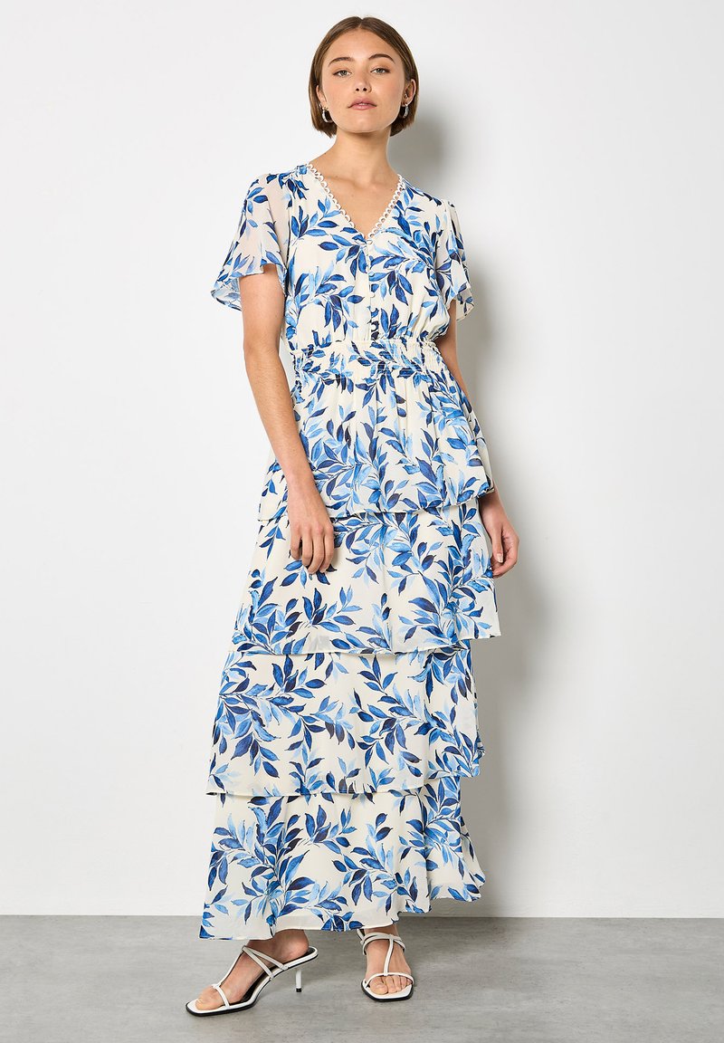 Robe longue en tissu léger avec imprimé feuilles bleues, encolure en V, manches courtes, taille élastique et ourlet à volants superposés. Portée avec des sandales à lanières.