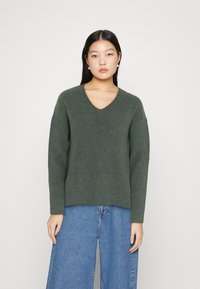 Pulover tricotat ribbed, de culoare verde închis, cu decolteu în V adânc și mâneci lungi, asortat cu blugi larghi din denim albastru. Design minimal, fără modele.