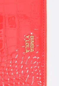 Czerwony portfel z lakierowanej skóry, o fakturze krokodylej, z wyraźnie widocznym złotym logo "BIMBA Y LOLA". Widać zamek błyskawiczny.