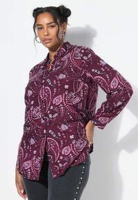 Vrouw met donker gevlochten haar, gekleed in een paarse button-up shirt met een paisleypatroon en donkere broek met zijperlenversieringen.