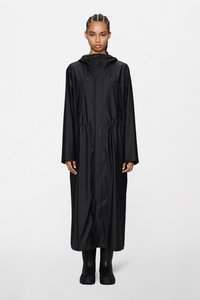 Γυναίκα που φοράει μακρύ μαύρο αδιάβροχο με κουκούλα και μπροστινές τσέπες, καθώς και μαύρα μποτάκια στον αστράγαλο, όρθια μπροστά σε λευκό, απλό φόντο.