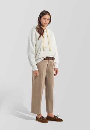 Mujer de pie con una sudadera blanca con capucha y cordones amarillos, pantalones beige de pierna ancha, cinturón marrón y mocasines marrones sobre un fondo gris claro.