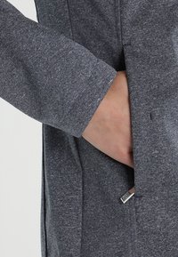 Šedá látková bunda s texturovaným povrchem, s boční kapsou a stříbrným kovovým prvkem. Rukáv je mírně ohrnutý.