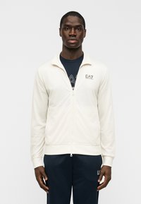 Ανοιχτό κρεμ ζακέτα με φερμουάρ, κατασκευασμένη από απαλό ύφασμα, με γιακά, πλαϊνές τσέπες και διακριτικό λογότυπο EA7 σε σκουρό καφέ στο στήθος.