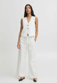 Chaleco blanco sin mangas con escote en V, que presenta dos botones dorados. Combinado con pantalones anchos blancos y bailarinas negras de punta.