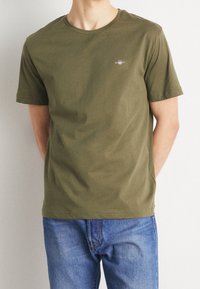 T-shirt in cotone verde oliva con colletto rotondo, maniche corte e un logo discreto sul petto; abbinato a jeans in denim blu.