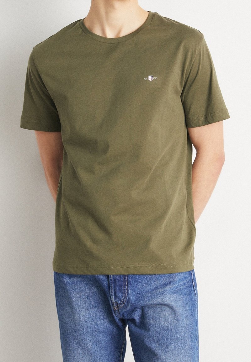 T-shirt in cotone verde oliva con colletto rotondo, maniche corte e un logo discreto sul petto; abbinato a jeans in denim blu.