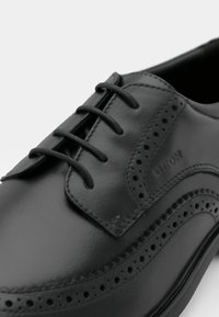 Chaussure en cuir noir avec des perforations, des lacets et une surface lisse. Logo de la marque 'GEOX' embossé sur le côté.