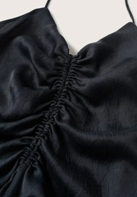 Musta satiinivaate, jossa on rypytetty yksityiskohta keskellä. Kankaassa on hieman ryppyinen rakenne ja ohuet säädettävät hihnat.