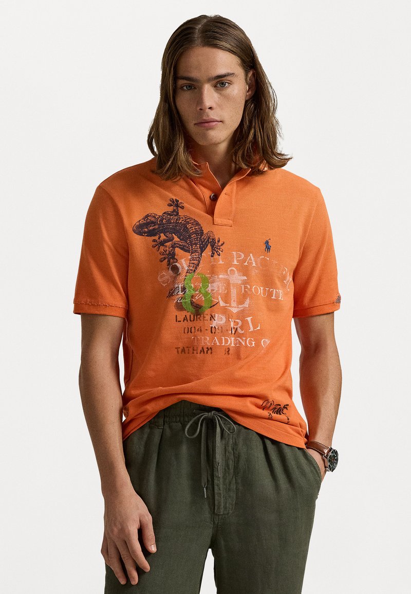 Oranje polo shirt met een knoopsluiting op de kraag, voorzien van een hagedisgrafiek en tekstdetails. Gecombineerd met ruime groene broek.