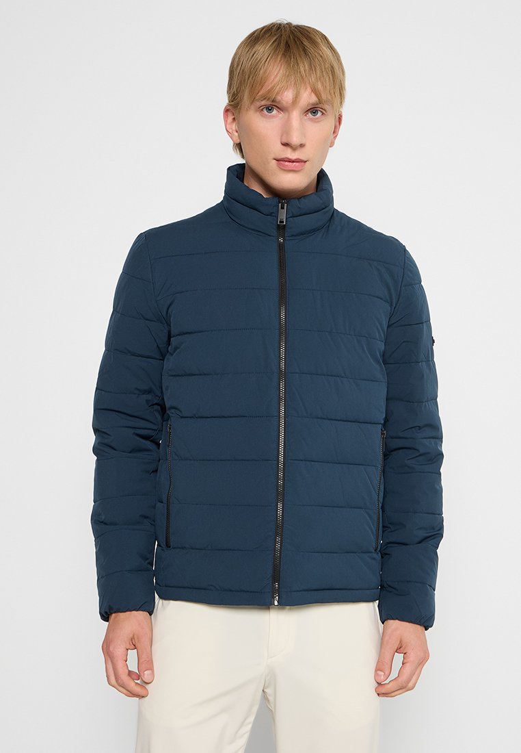 Marineblauwe gewatteerde jas met een hoge kraag, voorritssluiting en zijzakken. Voorzien van horizontale stiksels voor extra textuur.