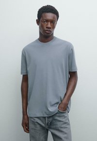 Šedé tričko s krátkým rukávem, kulatým výstřihem a jemnou texturou, v kombinaci se světle šedými džíny. Hladký, bezešvý design bez viditelných log.