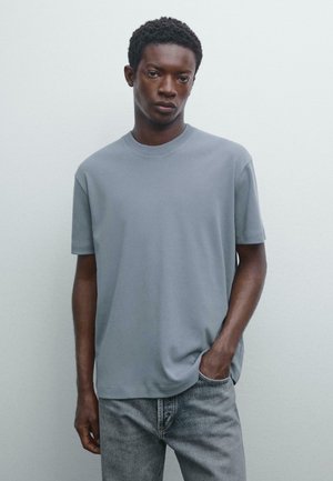 T-shirt gris à manches courtes avec un col rond et une texture douce, associé à un jean gris clair. Design lisse et sans couture sans logos visibles.
