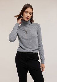 Hellgrauer Rollkragenpullover aus einem weichen, strukturierten Stoff, kombiniert mit schwarzen Hosen. Inklusive einer langen Halskette in Silberton.
