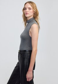 Šedý bezrukávový rolák vyrobený z měkkého materiálu, spárovaný s černými koženými kalhotami s vysokým pasem a černým opaskem se zlatým akcentem.