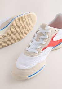 Fehér sportcipő bézs színű velúr részletekkel és piros logóval. Texturált gumitalppal és fehér fűzőkkel rendelkezik. Kék részlet a talp szélén.