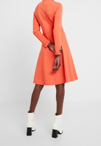 Långärmad klänning i knä längd i klart orange tyg med en figurnära överdel och utsvängd kjol. Matchad med vita ankelboots och kraftiga klackar.