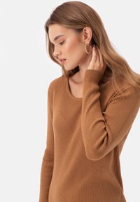 Brązowy sweter prążkowany z okrągłym dekoltem, długimi rękawami i teksturowanym materiałem. Modelka nosi złote kolczyki w kształcie kół i dotyka swojego włosa.