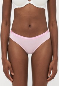 Woman wearing light pink cotton bikini-style underwear and white bra against plain background, showing torso and upper legs.