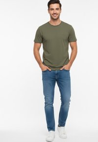 Camiseta de manga corta verde oliva con cuello redondo, combinada con jeans de mezclilla azul. La modelo lleva zapatillas blancas y tiene las manos en los bolsillos.